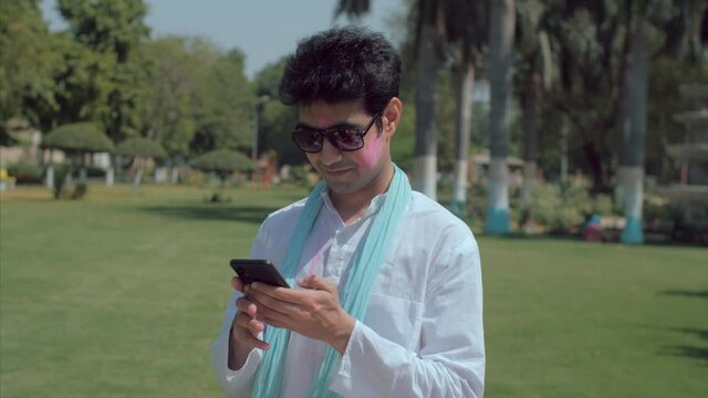 Young male with Gulal on his face using his mobile on Holi festival celebration. The Indian festival of colors party - Charming male in Kurta-Pajama standing outdoors with Abeer on face