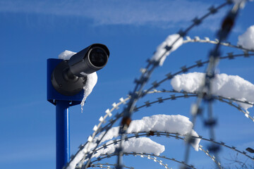 Barbed wire and security camera covered with snow against a blue sky.