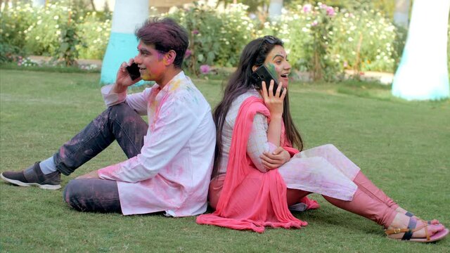 Two youngsters talking on their mobiles with their backs against each other - Holi Day. Cheerful male and female sitting and relaxing outdoors in the park after Holi party