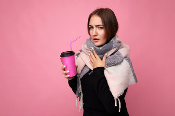 Photo shot of beautiful young brunette woman wearing black sweater and warm scarf isolated over pink background holding paper coffee cup for mockup drinking hot beverage and having cold