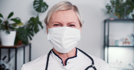 Portraif of happy senior Caucasian female doctor posing at office in protective face mask, white...