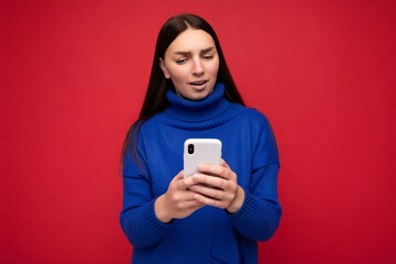 Thoughtful attractive good looking young brunette woman wearing stylish blue warm sweater poising isolated on red background with empty space holding in hand and using mobile phone reading news online