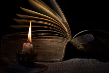 A candle burns in the dark. In the background there is an old book with open pages. The concept of...