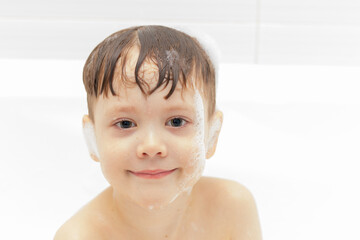 a preschool boy washes in a light white bathtub and plays with lather. close-up portrait