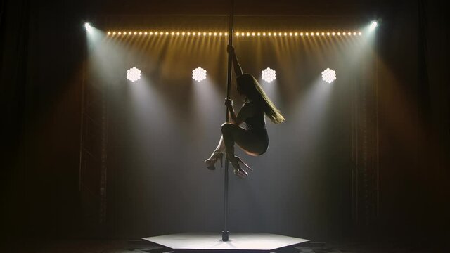 Professional pole dancer performs tricks while spinning on pole. Stripper with long hair erotically moves on pole in dark studio with bright lights. Erotic dance, strip show. Silhouette. Slow motion.