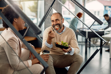 Happy businessman eating lunch while relaxing on staircase and talking to female co-worker.