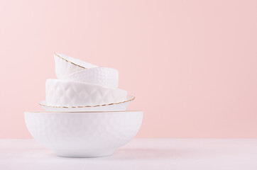 White ceramic dishes with thin gold border, different tracery form on white wood table in delicate soft light pastel pink interior.