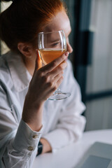 Close-up of beautiful redhead young woman holding glass with champagne near face, looking away. Portrait of pretty female with glass of wine.