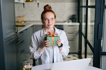Portrait of happy charming redhead young woman holding present gift box during video call via laptop webcam. Concept of leisure activity red-haired female at home during self-isolation.