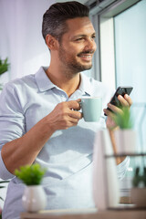 cheerful man stood by window holding coffee and smartphone