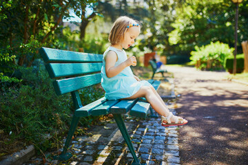 Adorable toddler girl sitting on the bench outdoors and having snack on a sunny summer day