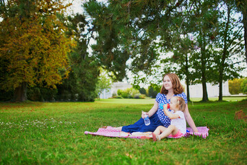 Woman and adorable toddler girl sitting on the ground and having picnic in summer park. Mother and daughter enjoying sunny day together. Outdoor activites for families