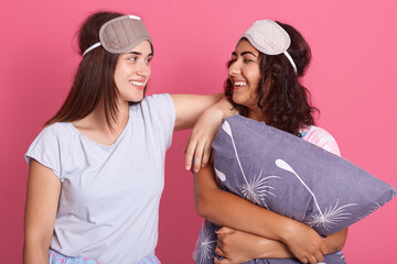 Two European young girls in pajamas and sleep masks isolated over pink background, looking at each other with happy smile, dark haired females on pajamas party.