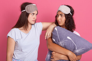 Two good looking attractive young brunettes with pillow, going to sleep, wearing pajamas and sleeping masks on heads, standing isolated over pink background, being serious, looking at each others.