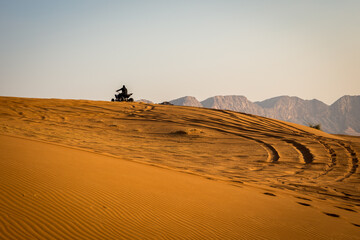 Silhouette of a quad driving up the sand dune in the desert, with tire tracks behind and mountains in the background, sunset, Fossil Rock, Sharjah, United Arab Emirates.