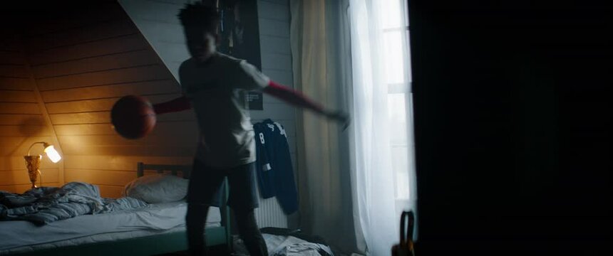 WIDE HANDHELD African American Black kid teenager boy playing with basketball in his attic bedroom at home. Shot with 2x anamorphic lens