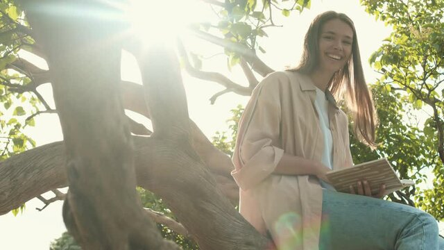 Medium long POV of happy beautiful Caucasian woman sitting on tree in sunny summer park, reading book, then waving hand, smiling and looking on camera