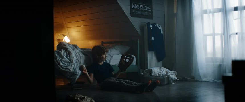 WIDE cute little Caucasian kid sitting in his bedroom, playing baseball catch with himself at home. Shot with 2x anamorphic lens