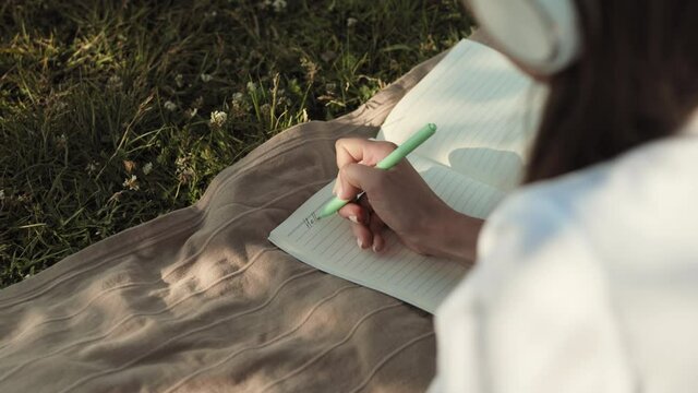 Over shoulder of unrecognizable person lying on blanket over green lawn, writing hello in journal with pen