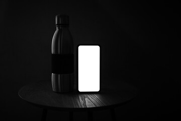 Dark photo in black and white of smartphone with mockup and steel reusable thermo bottle on wooden table against black background.