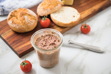 Grobe Leberwurst im Glas aus Vorrat mit Brötchen, Butter und Tomaten als Vesper Brotzeit auf Holzbrett und Marmor Hintergrund hell