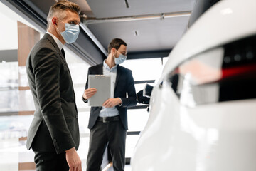 Mid white manager in face mask showing car to customer in showroom