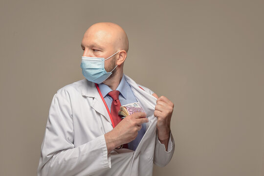 Bald Male Doctor Putting Wad Of Cash In His Hand Into His Jacket Inner Pocket And Looking Over His Shoulder . Corruption, Bribery And Kick Back Concept.