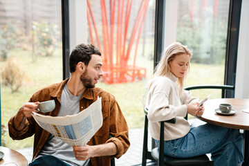White man looking at blonde woman while reading newspaper in cafe