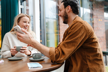 Smiling man and woman talking and using cellphone while drinking coffee