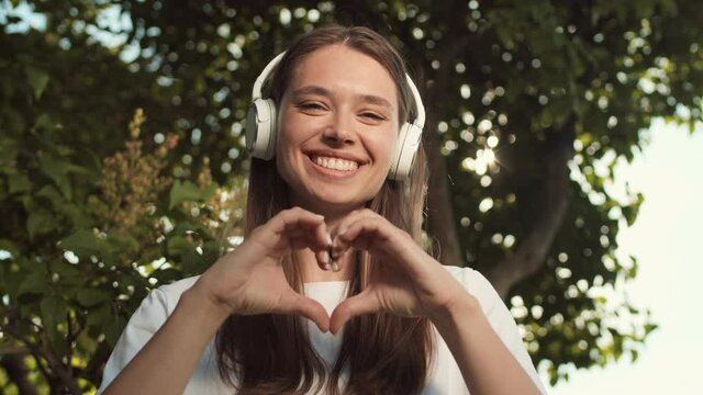 Tilting up POV of young happy Caucasian woman wearing over-ear headphones, showing heart sign with hands, smiling and looking on camera