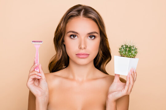 Photo of young serious beautiful girl wear no clothes hold cactus and razor depilation isolated on beige color background