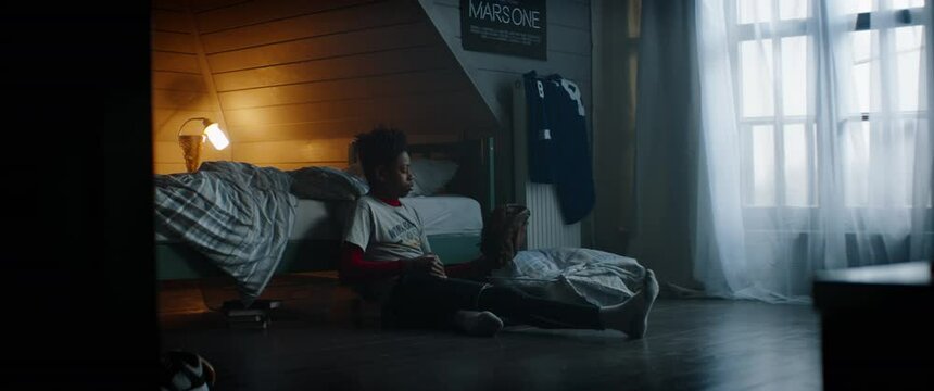 WIDE Bored African American Black kid teenager boy playing baseball catch with himself in his attic bedroom at home. Shot with 2x anamorphic lens