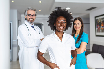 Obraz premium A multi-ethnic group of three doctors and nurses standing in a hospital corridor, wearing scrubs and coats. The team of healthcare workers are staring at the camera and smiling