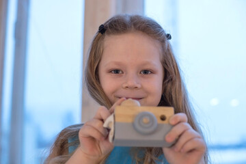 A little girl with long blonde hair looks straight and wants to take a picture of someone.
