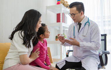 Young Asian man in glasses with stethoscope explain about medical equipment to little girl sitting with mother in office. Kid and family healthcare concept