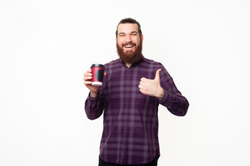 Photo of cheerful young bearded man in casual showing thumb up and holding cup of coffee to go