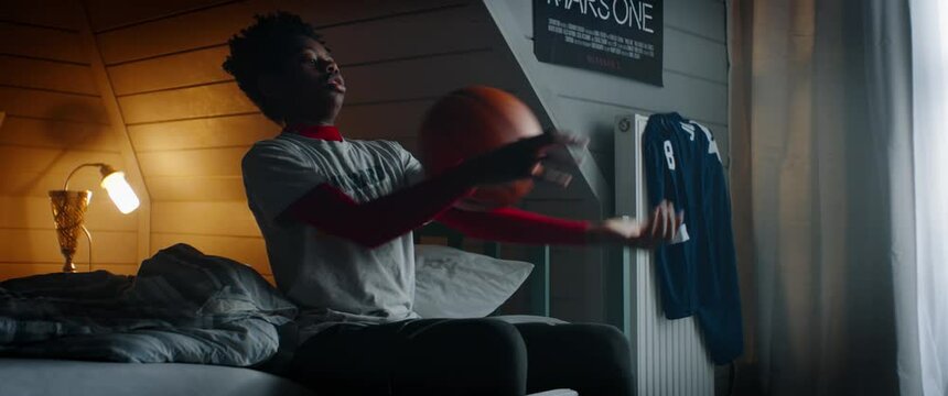 WIDE HANDHELD African American Black kid teenager boy playing with basketball in his attic bedroom at home. Shot with 2x anamorphic lens
