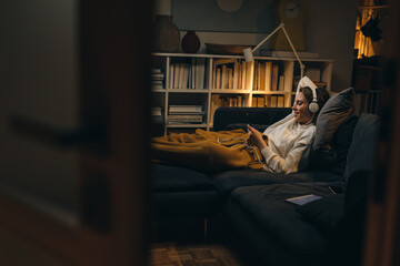 woman relaxing on sofa at her home and listening to a music on headphones. night moody ambience