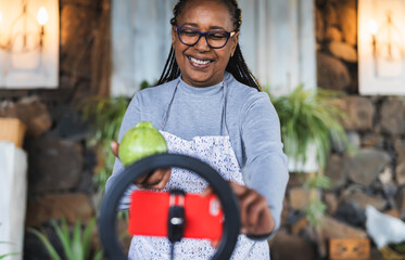 African senior woman preparing food recipe while streaming online with mobile smartphone cam for...