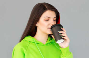 Fototapeta premium Studio shot of girl with holds paper cup of hot coffee, wears green jumper, grey background. Beautiful young woman enjoys aromatic cappuccino.