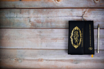 Picture of Quran with pen against wooden board background. The big Arabic word that mentions the Quran is by the method of reading the letters and the method of correct pronunciation. Copyspace
