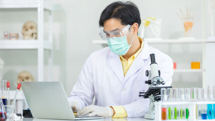 Asian man scientist wearing equipment mask and Protective glasses for working to computer laptop microscope while working on medical research in science laboratory. Concept research  provent 