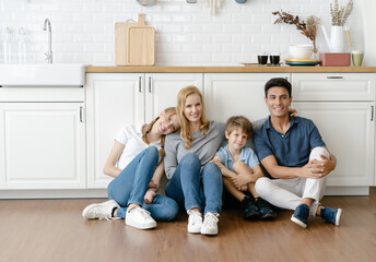 Portrait of Happy Caucasian family with teenage daughter and little son sitting on warm wooden floor in modern kitchen and looking at camera. Family enjoying weekend at home together.
