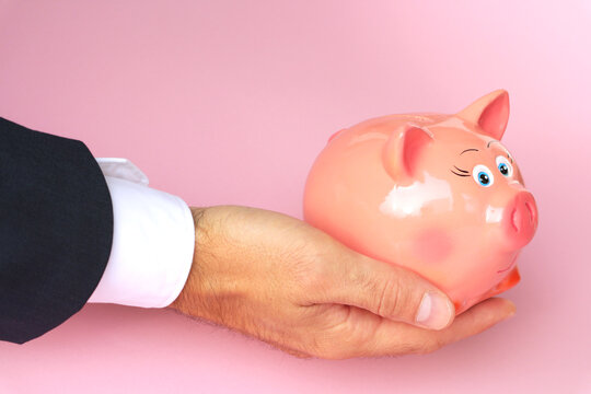 A man holds a piggy bank in his hand on a pink background.