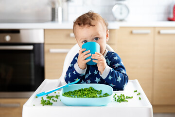Cute baby eating grean peas in high chair drinking with cup