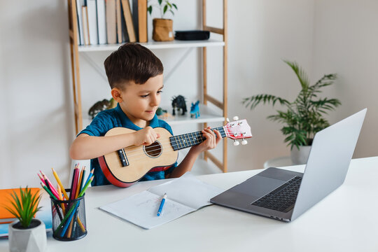 Kid looking online video lesson about soprano ukulele via laptop. Elementary school boy learning play guitar at leisure. Concept of early childhood education, e-learning, talent and music hobby