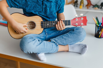 Close-up kid playing soprano ukulele sitting on desk. Preschool boy learning guitar at leisure. Concept of early childhood education and music hobby