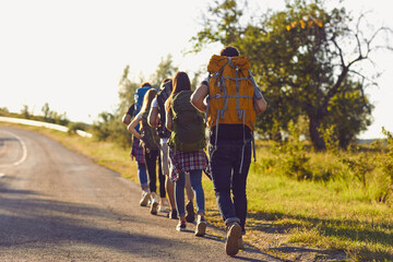 Obraz premium Group of young hikers walk in a row along the road with backpacks on their shoulders.