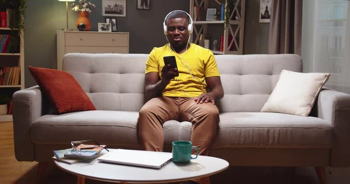 Good-looking African American guy sitting on sofa and texting friend on smartphone at home. Young guy in headphones listening to music, chilling and having fun in evening. Entertainment concept.