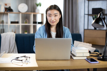 Beautiful asian girl student with long dark hair working indoors at home on wireless laptop while sitting at desk. Female freelancer in denim shirt using computer for remote work.